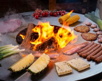 Feuerplatte mit Burgern, Würstchen, Gemüse und Ananas rund um ein Holzfeuer